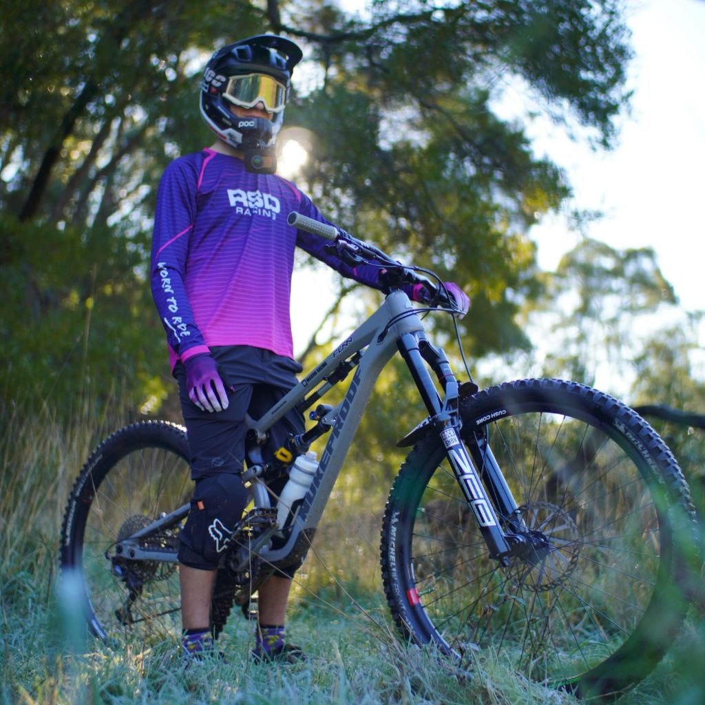 Rider in a purple RSD Racing jersey and full face helmet standing with a bike in long grass at sunset